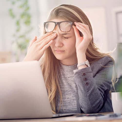 A tired woman sitting in front of her computer while removing her glasses